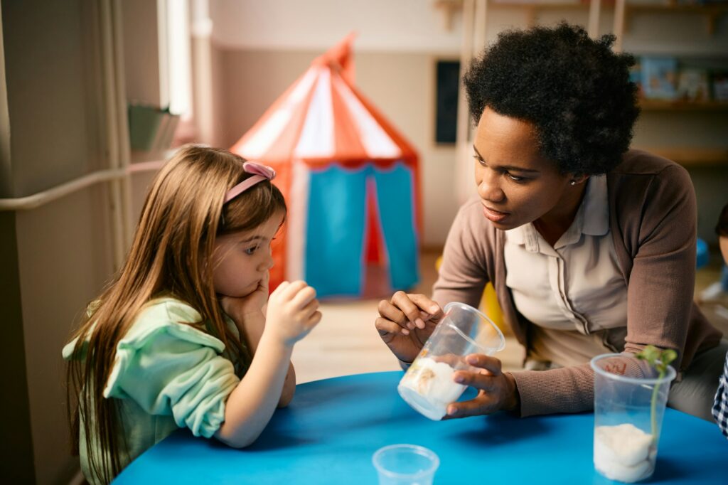 African American preschool teacher assisting little girl with planting seeds during biology class.